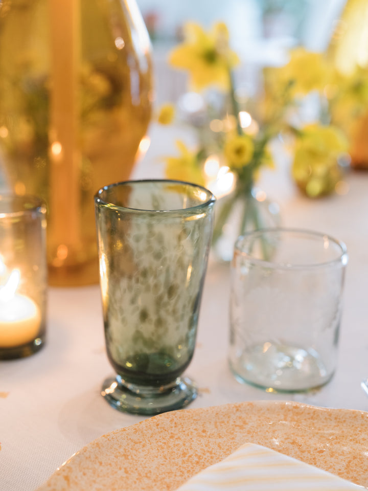 A styled tabletop scene with two drinking glasses, one smoky green and one clear, alongside a lit candle, a large gold vase, and blurred yellow flowers in the background.