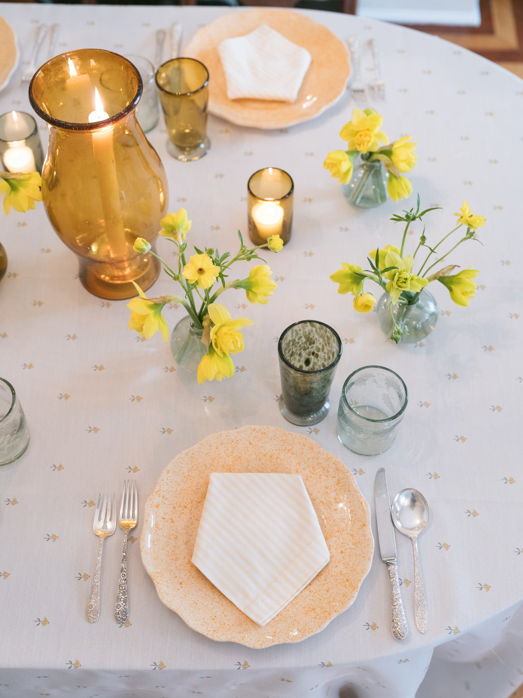 A styled round table setting with a white tablecloth, beige patterned plates, silver cutlery, amber glass vases, and small yellow flower arrangements with lit candles.