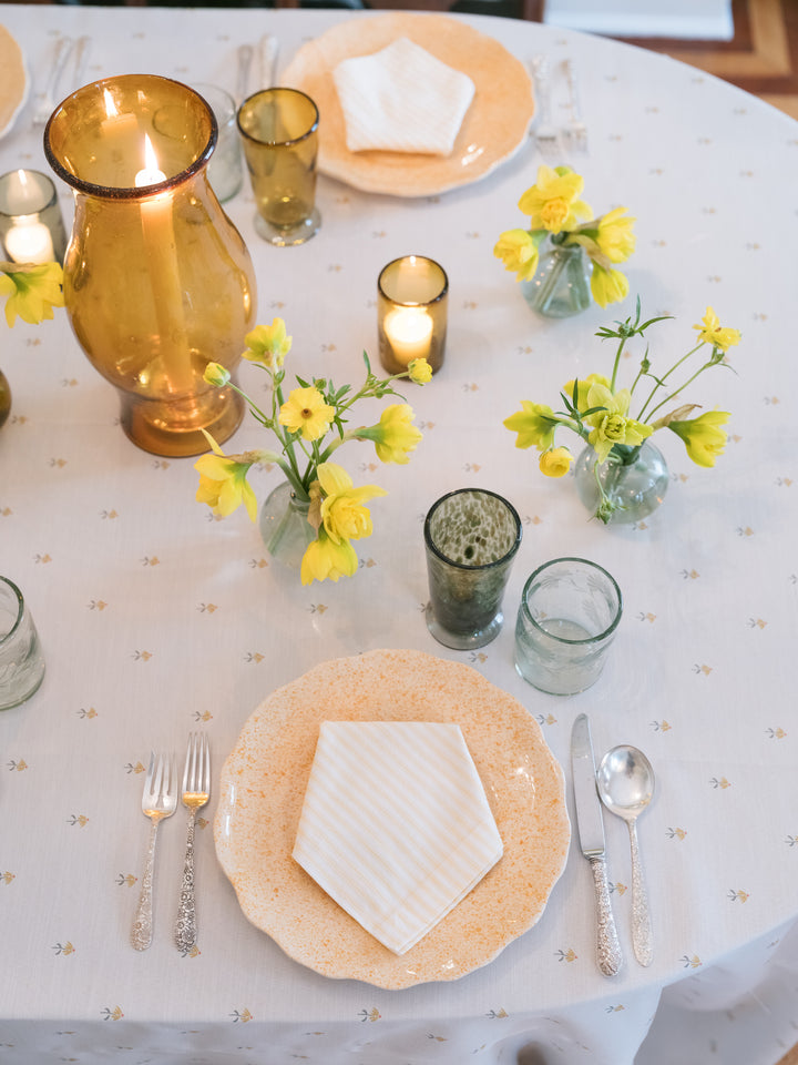 A styled round table setting with a white tablecloth, beige patterned plates, silver cutlery, amber glass vases, and small yellow flower arrangements with lit candles.
