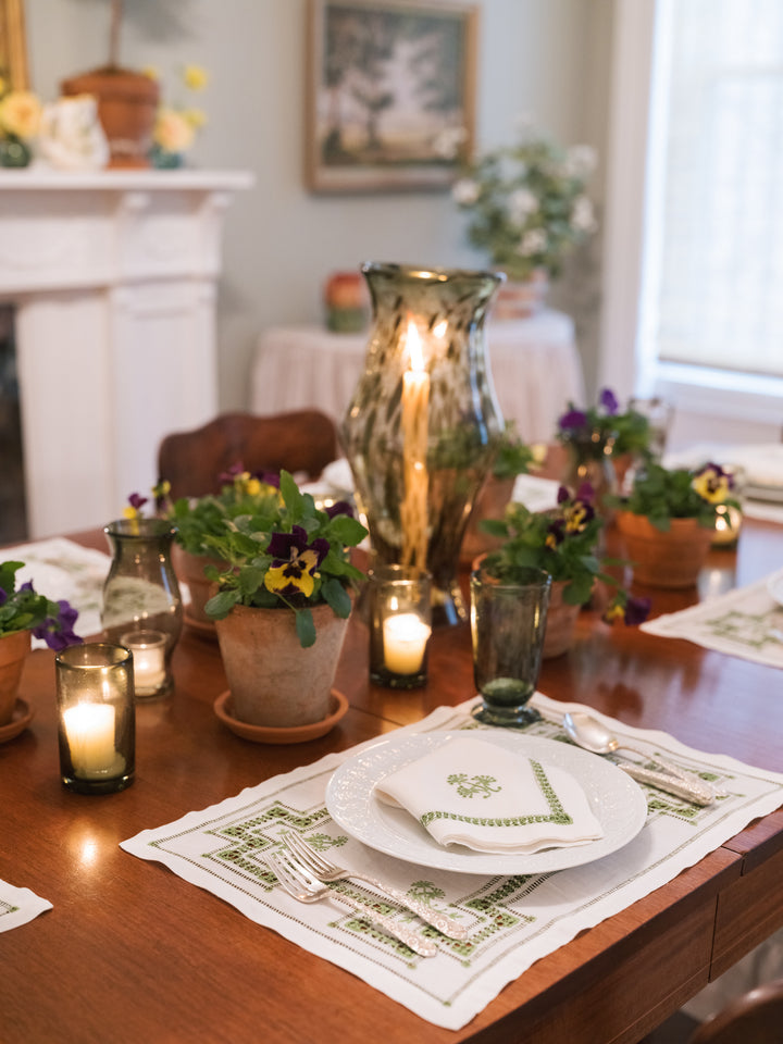 A styled dining table setting in a warm indoor room, featuring embroidered white placemats with green geometric borders, white plates, folded napkins with green embroidery, glass candle holders, and small potted pansies.