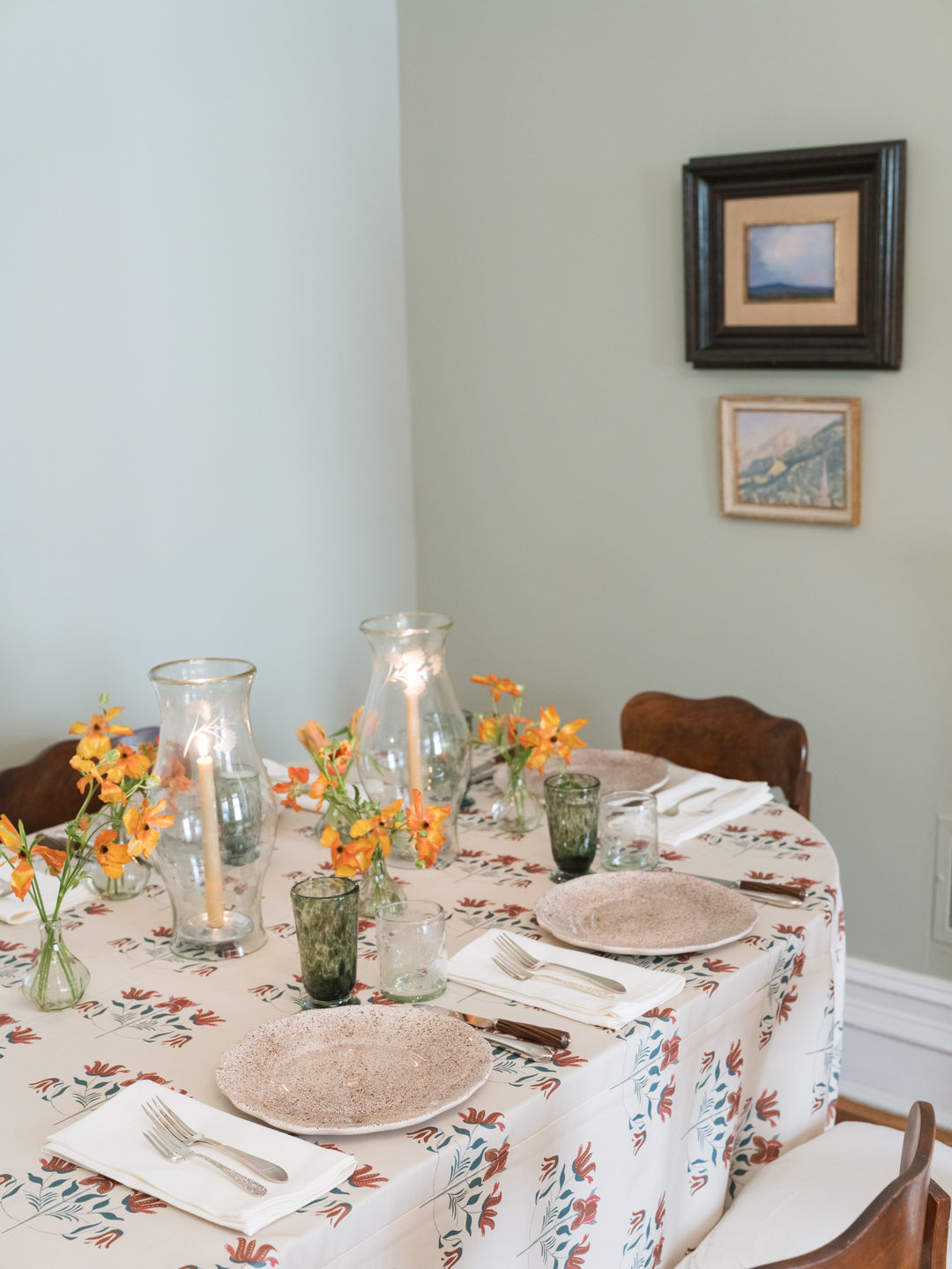A dining table set for four with a floral tablecloth, textured plates, green glass tumblers, and orange flower arrangements in a softly lit room with pale green walls and framed art.