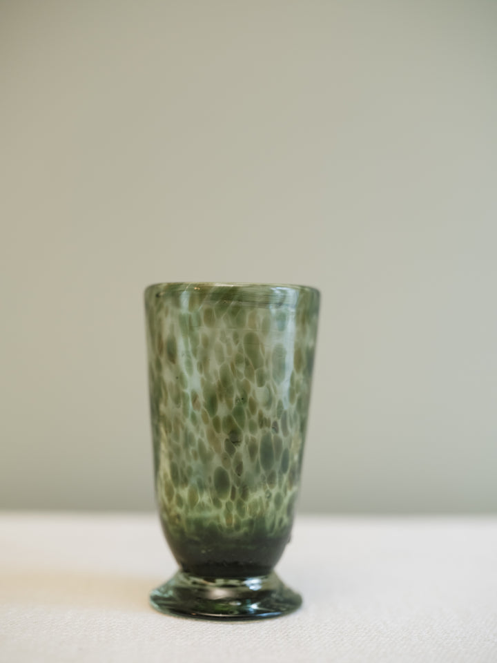A tall, slender green glass tumbler with a textured, marbled pattern and a flared base, standing on a light surface against a neutral gray background.