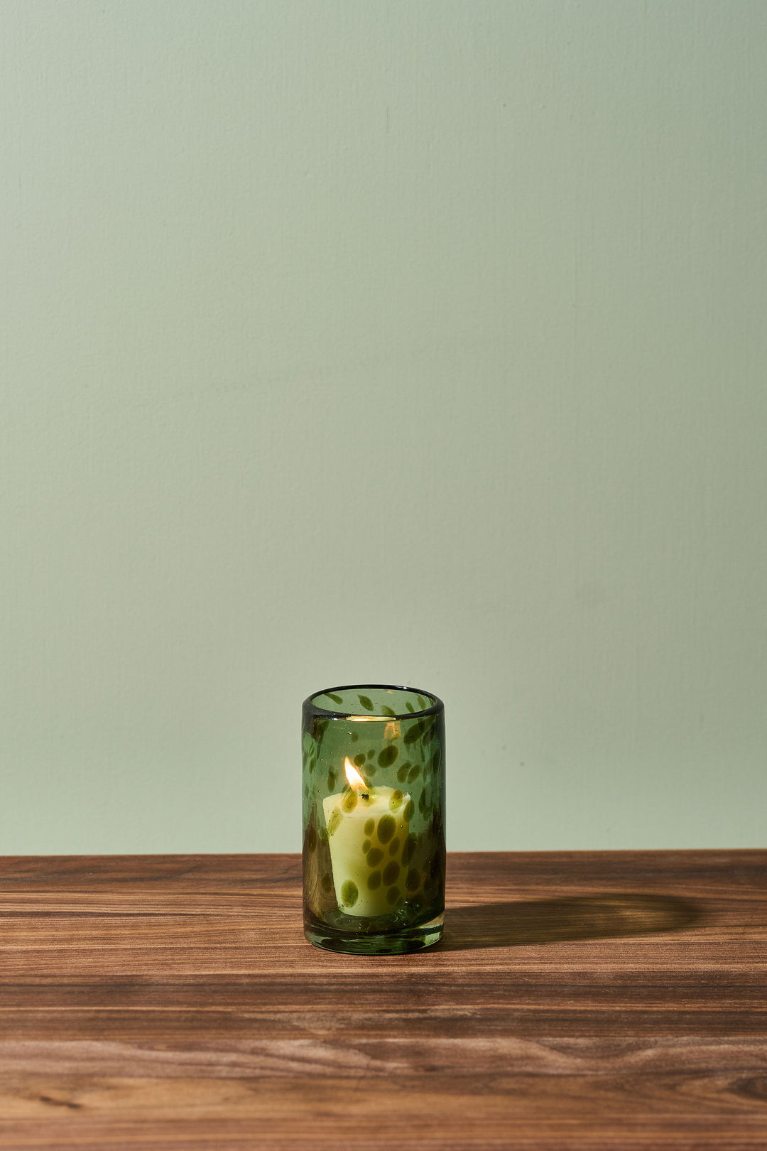 A small green glass candle holder with a lit candle inside, set on a wooden tabletop against a pale sage green wall.