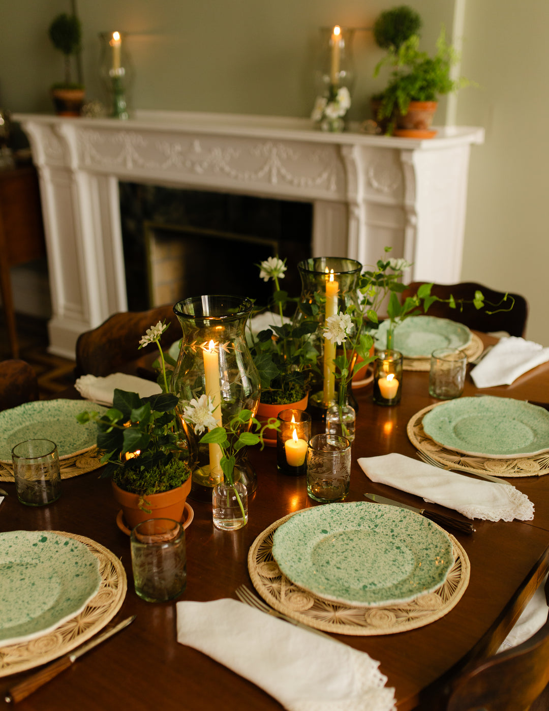 A styled dining table set for four with speckled sage green ceramic plates, woven placemats, white napkins, and botanical glass candle holders with lit candles, set in a warm room with a white fireplace in the background.