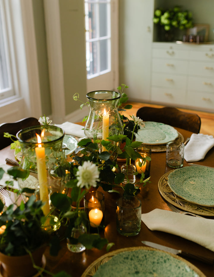 A styled dining table centerpiece with green glass candle holders, lit taper candles, white flowers, and green speckled plates in a warm, softly lit room.