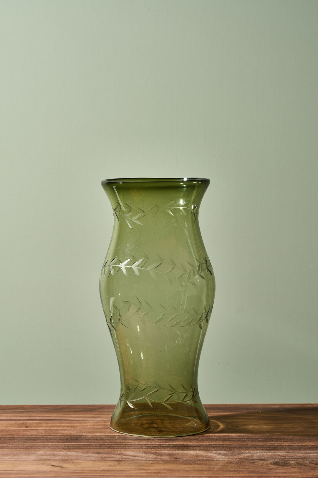 An olive green glass vase with a flared rim and etched leaf patterns, standing on a wooden surface against a light sage green background.