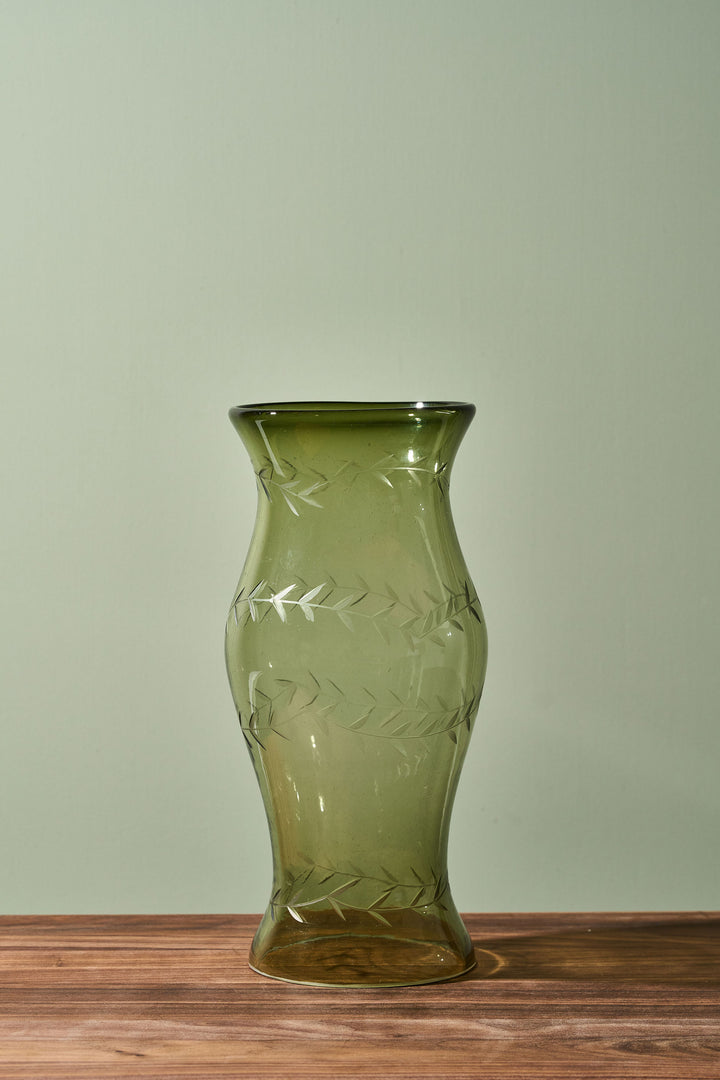 An olive green glass vase with a flared rim and etched leaf patterns, standing on a wooden surface against a light sage green background.