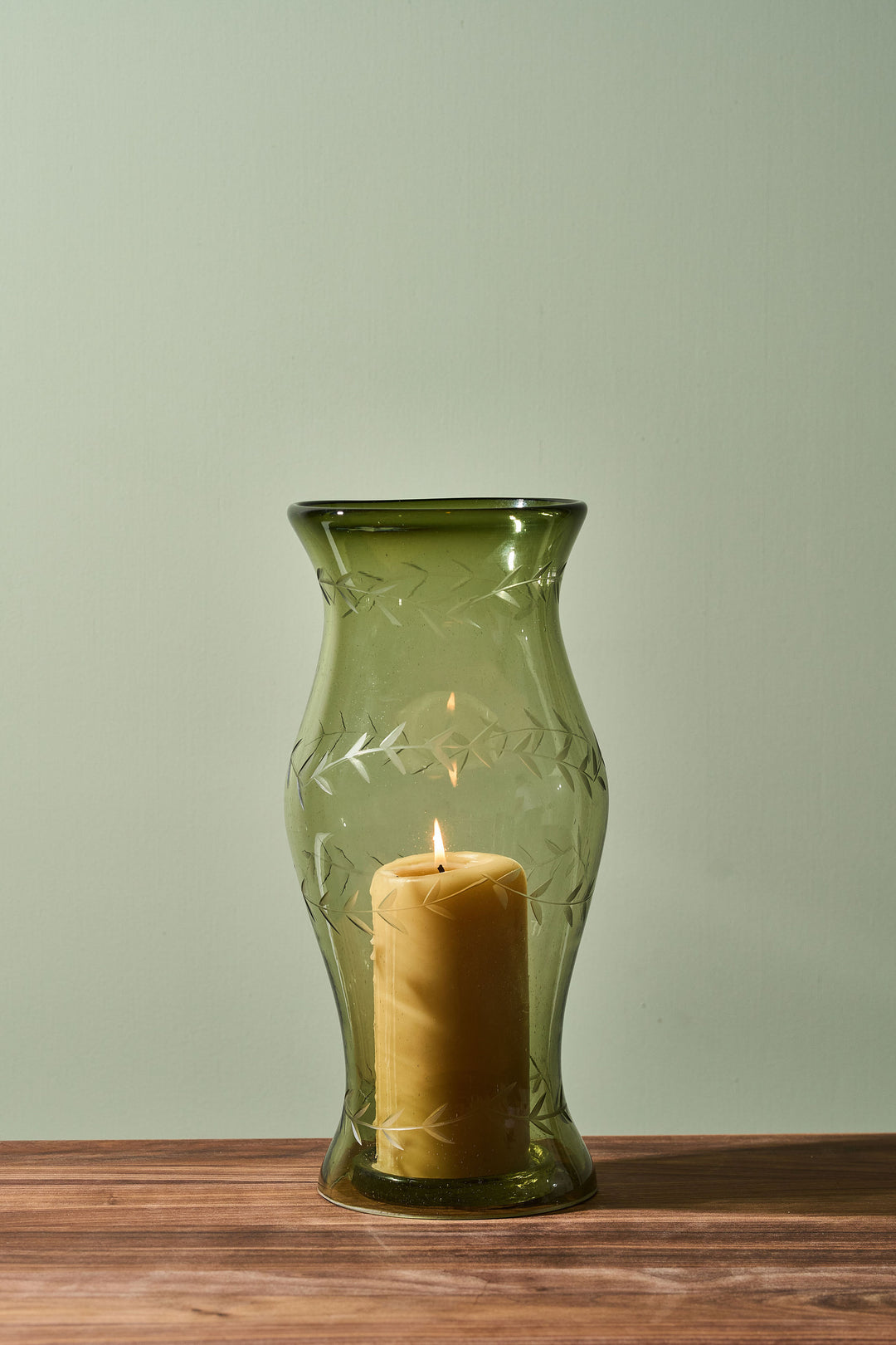 An olive green glass vase with etched leaf patterns, holding a lit yellow pillar candle, set on a wooden surface against a light sage green background.