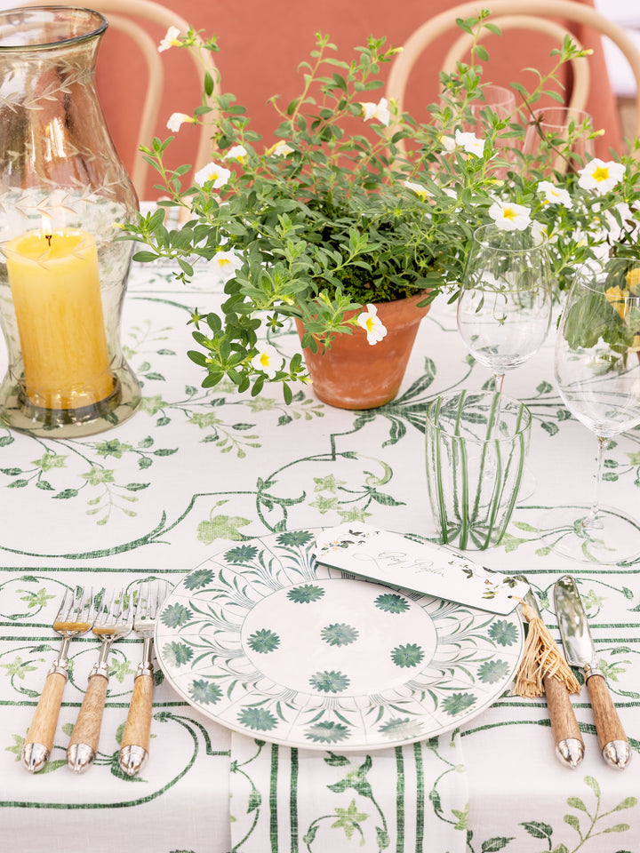 A styled dining table setting featuring a white tablecloth with a green botanical pattern, matching patterned plates, silver cutlery with light wood handles, a yellow candle in a glass vessel, and a potted green plant in the center.