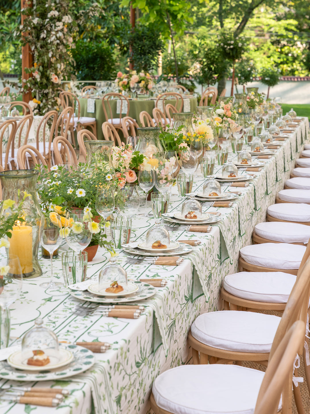 A long outdoor wedding reception table set with a green and white botanical print tablecloth, white plates, clear glass goblets, floral centerpieces with yellow and peach flowers, and light wood chairs with white seat cushions.