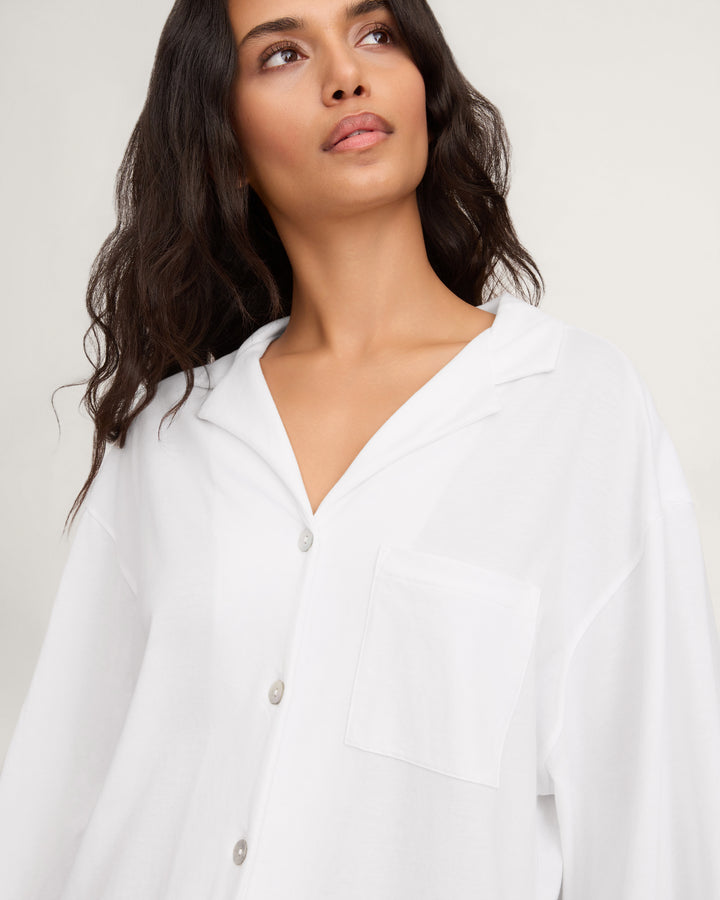 A woman wearing a white long-sleeve button-up shirt with a relaxed fit, a chest pocket, and a V-neckline, photographed against a plain light background.