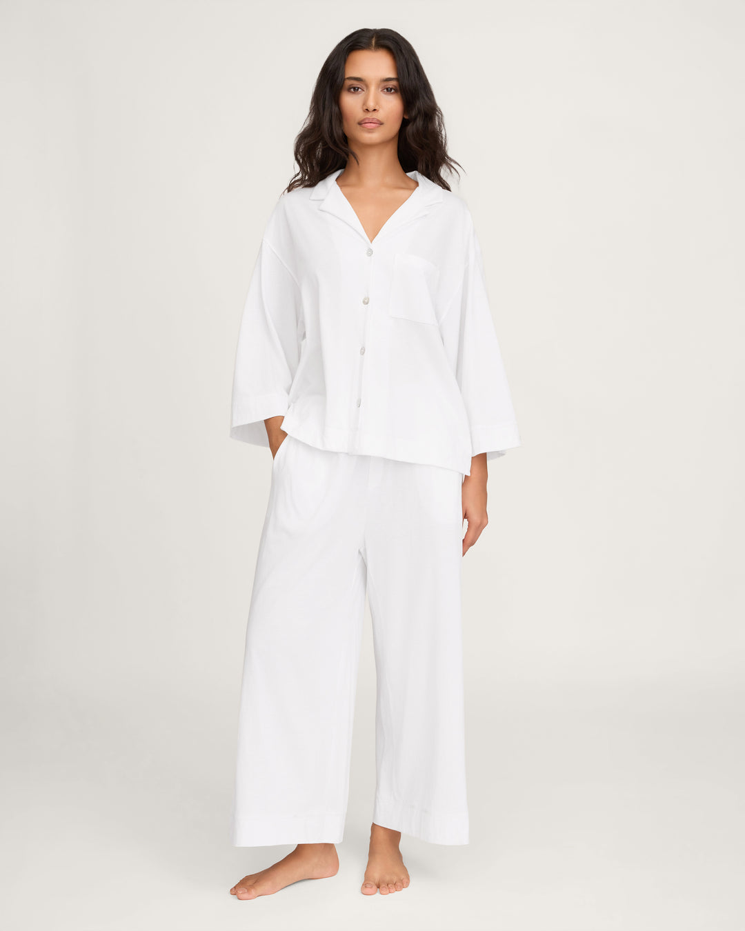 A woman modeling a white two-piece pajama set consisting of a button-front long-sleeve shirt and matching wide-leg pants, photographed against a plain light studio background.