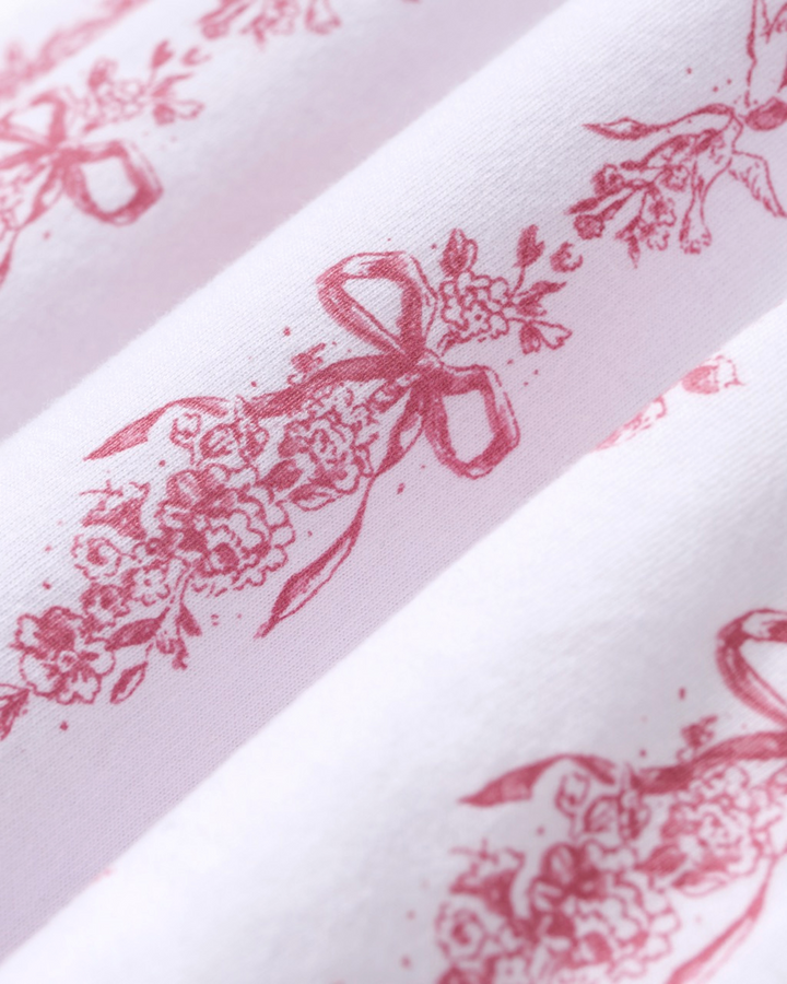 White fabric with a repeating dusty rose red floral and bow toile pattern, shown in soft diagonal folds.