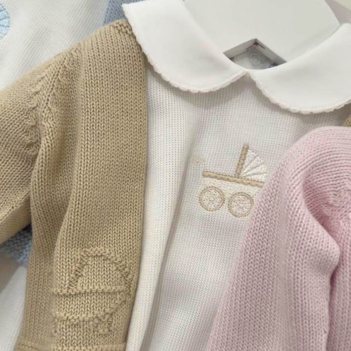 A close-up of a baby knit outfit featuring a cream-colored long-sleeve top with a white Peter Pan collar and a beige embroidered baby stroller motif, layered with a beige knit cardigan and a pink knit sleeve.