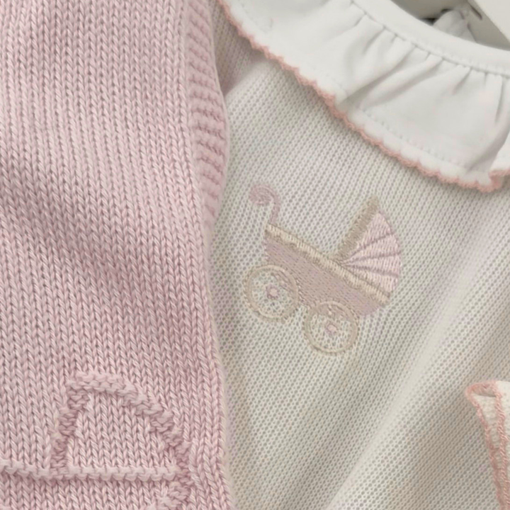 Close-up of a baby clothing set featuring a light pink knit vest and a cream-colored knit top with a ruffled collar and an embroidered baby stroller motif.