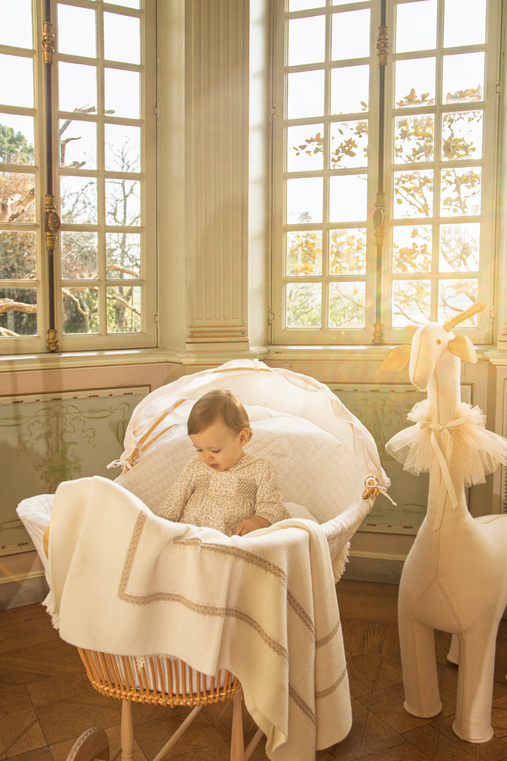 A baby sits in a woven bassinet draped with a cream blanket, placed in a sunlit room with tall windows. A cream stuffed giraffe toy stands beside the bassinet.