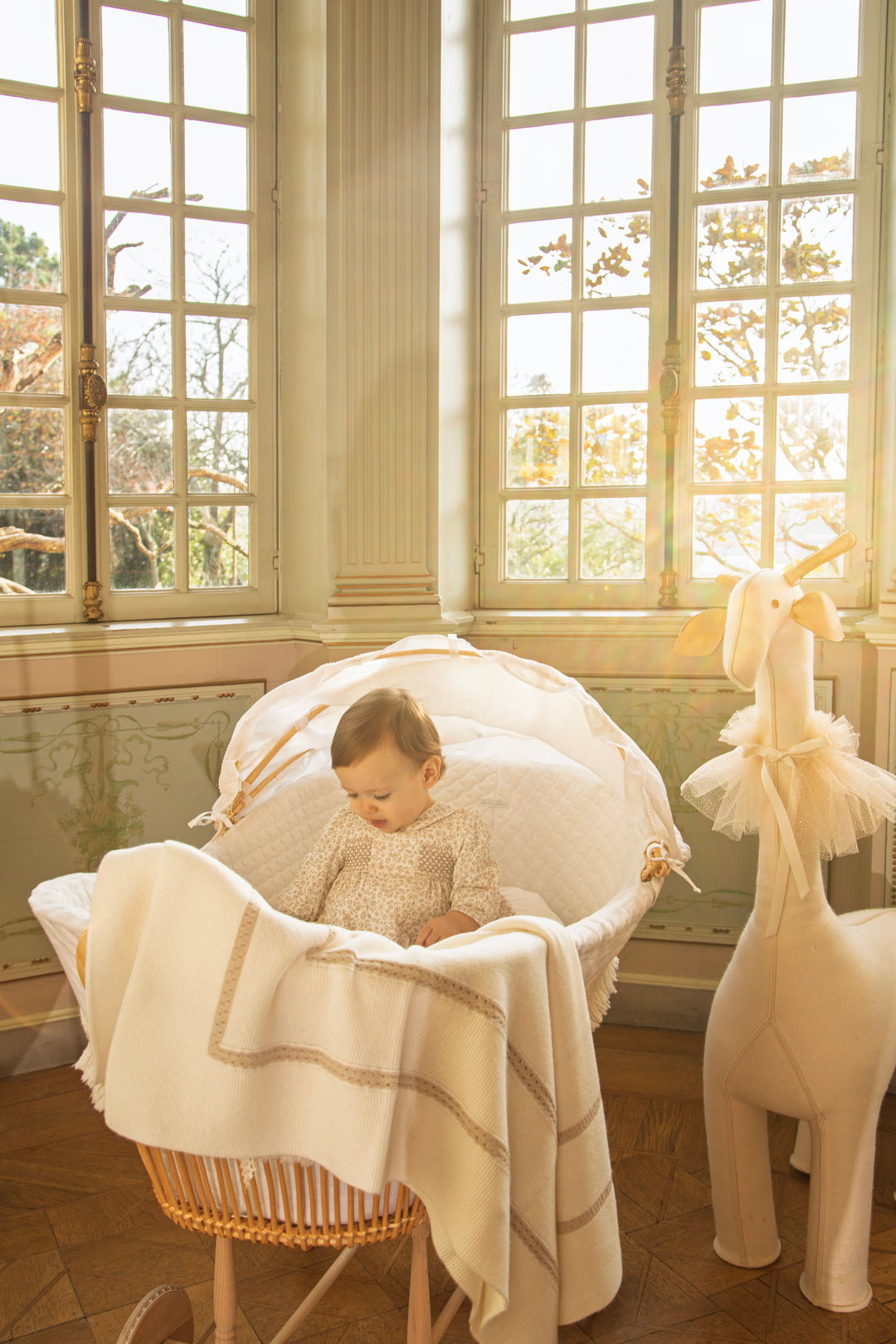 A baby sits in a woven bassinet draped with a cream blanket, placed in a sunlit room with tall windows. A cream stuffed giraffe toy stands beside the bassinet.