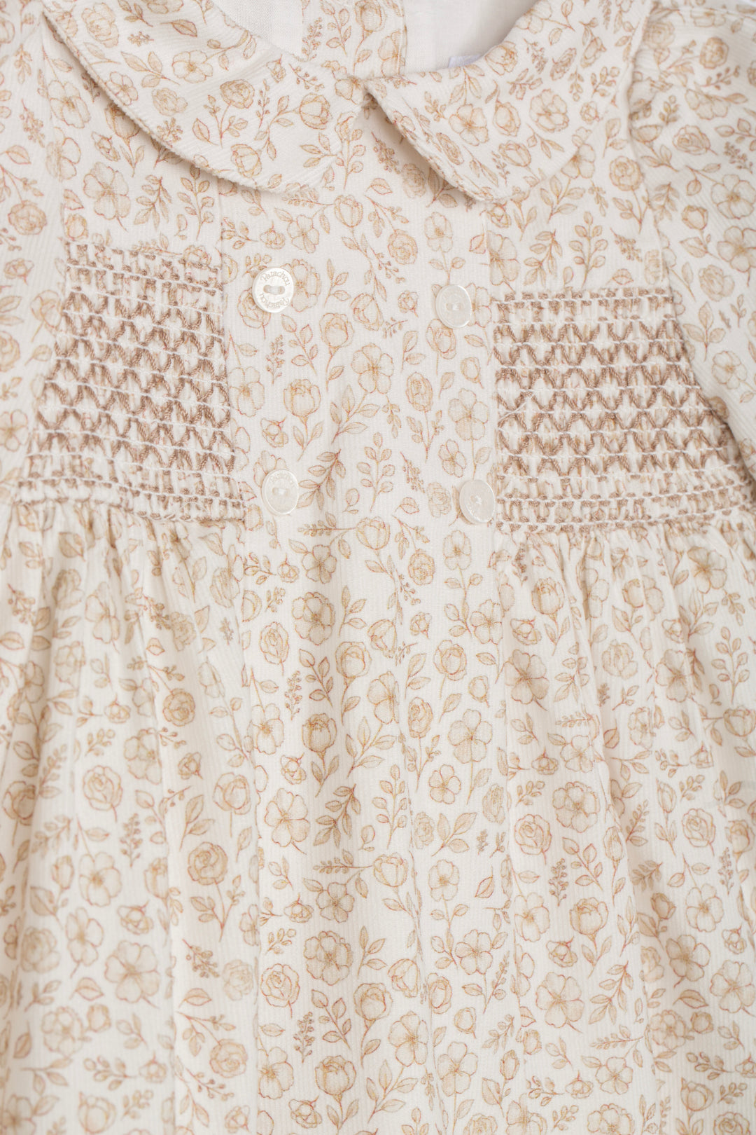Close-up of a baby dress featuring a beige and cream floral print, a Peter Pan collar, and smocked chest panels with a single white button.