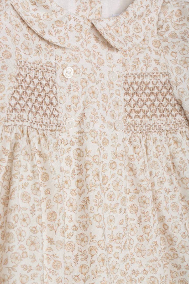 Close-up of a baby dress featuring a beige and cream floral print, a Peter Pan collar, and smocked chest panels with a single white button.