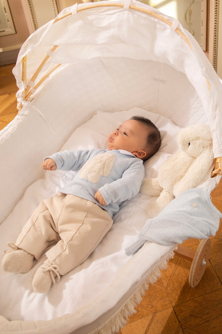 A baby wearing a light blue long-sleeve top and beige pants lies on their back inside a white bassinet with a wooden frame and a plush cream teddy bear beside them.