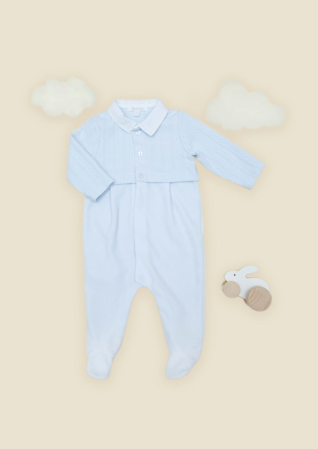A light blue long-sleeve baby footed romper with a buttoned collar, laid flat on a warm beige background with decorative white cloud shapes and a small wooden bunny toy.