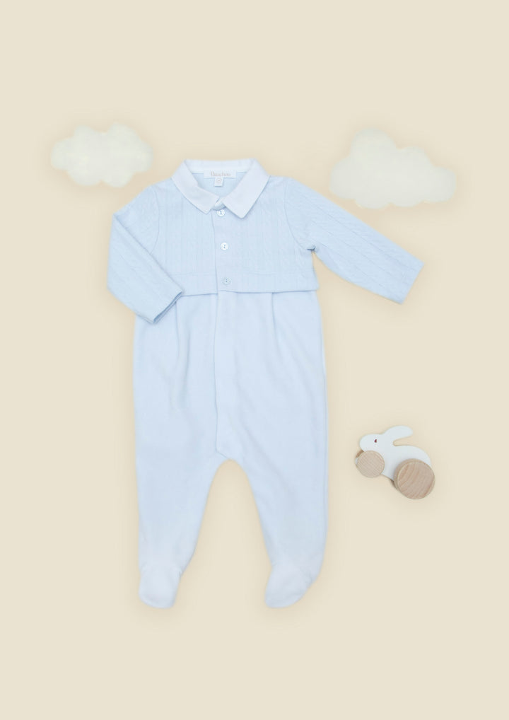 A light blue long-sleeve baby footed romper with a buttoned collar, laid flat on a warm beige background with decorative white cloud shapes and a small wooden bunny toy.