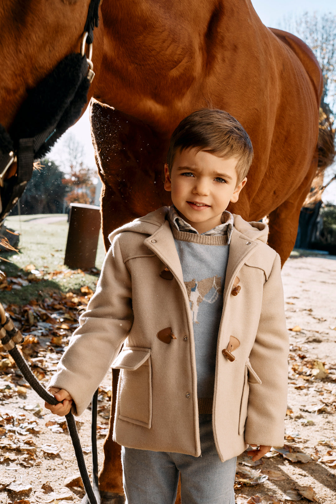 A young boy wearing a beige duffle coat over a gray sweater stands outdoors in front of a brown horse, holding a lead rope.