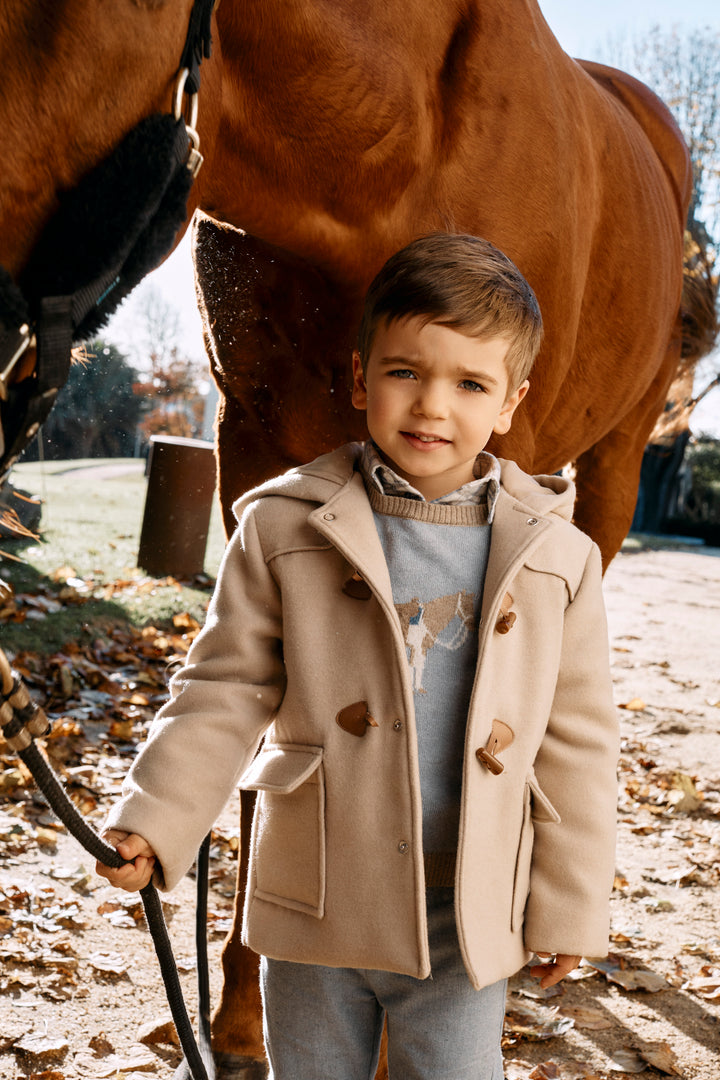 A young boy wearing a beige duffle coat over a gray sweater stands outdoors in front of a brown horse, holding a lead rope.
