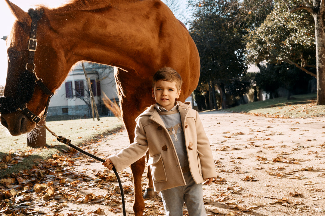 A young boy in a beige coat stands outdoors next to a brown horse, holding a black lead rope. The scene is set in a sunny, leaf-strewn yard with trees in the background.