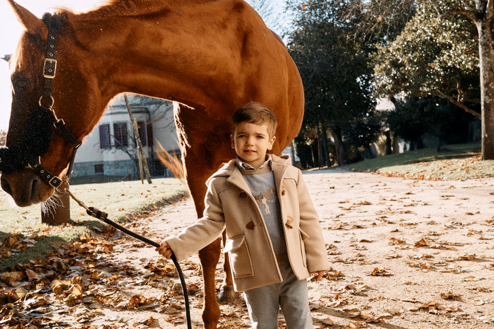A young boy in a beige coat stands outdoors next to a brown horse, holding a black lead rope. The scene is set in a sunny, leaf-strewn yard with trees in the background.