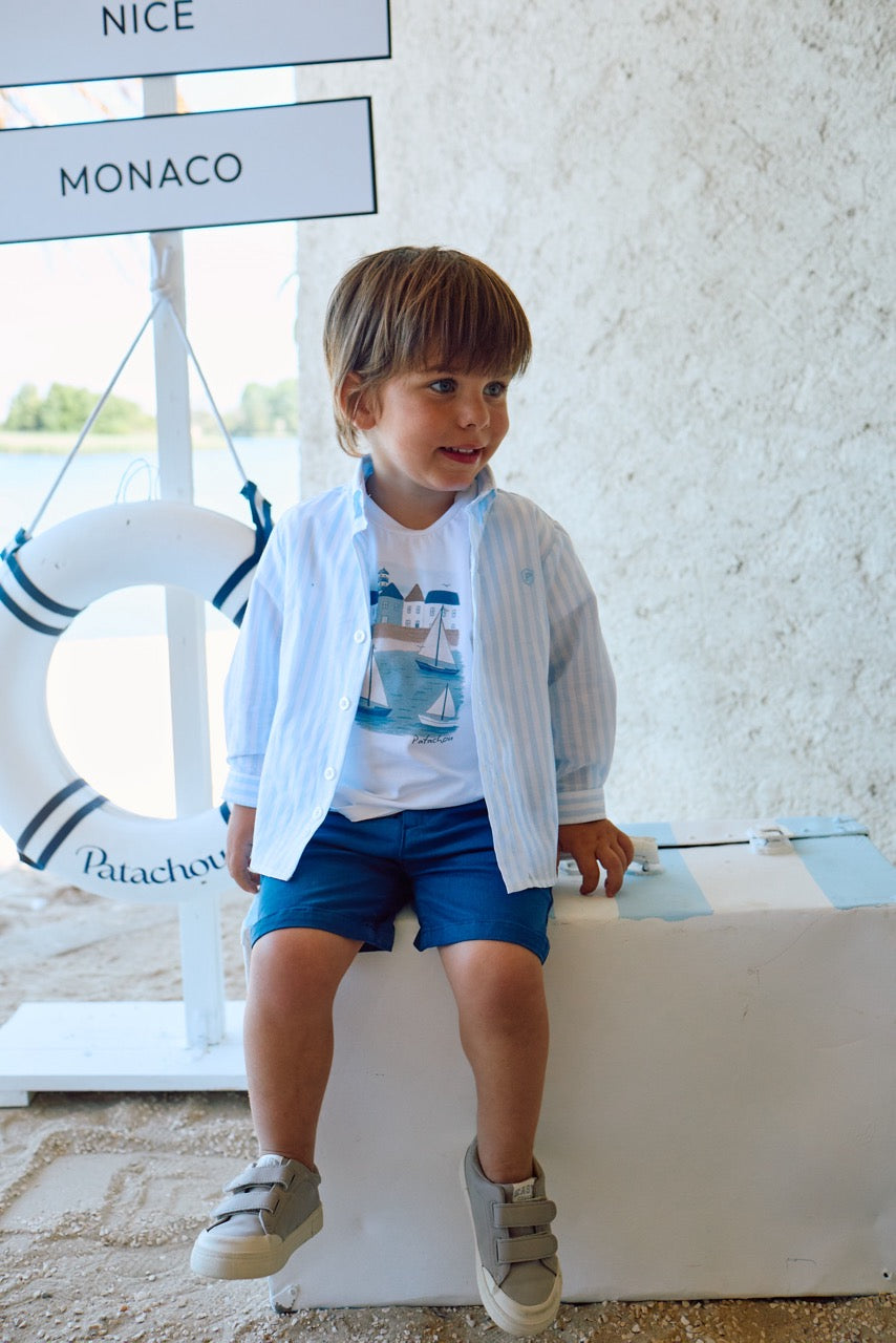Child wearing a light blue jacket over a white shirt with a sailboat design, sitting on a white ledge.