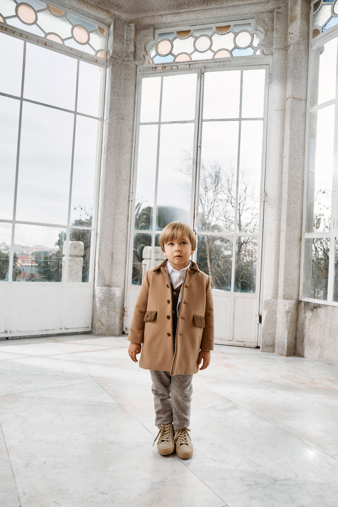 A young boy stands in a large, sunlit room with tall industrial windows and concrete pillars. He is wearing a camel brown coat over a dark shirt, gray trousers, and brown shoes.