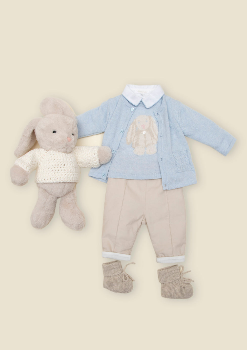 A baby clothing set laid flat on a warm beige background, including a light blue cardigan with a white collar, beige pants with white cuffs, beige booties, and a plush gray bunny toy wearing a white knit sweater.