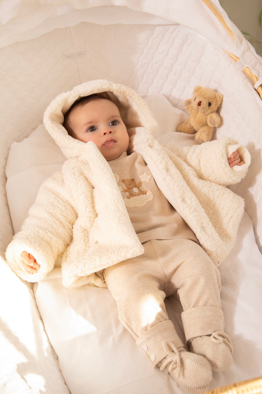 A baby lying in a soft cream-colored bassinet, wearing a matching hooded fleece jacket, one-piece outfit, and booties, with a small teddy bear toy beside them.