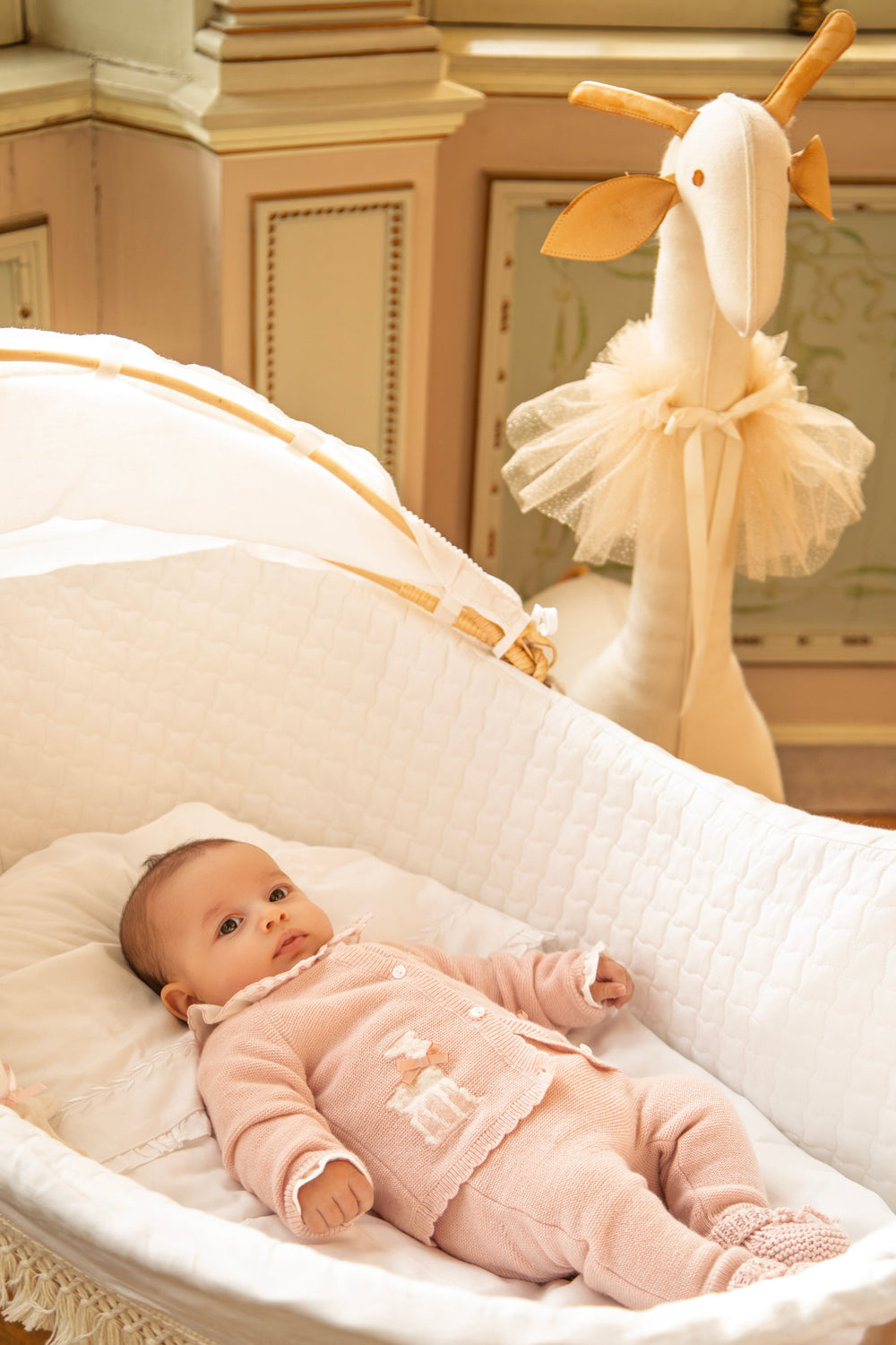 A baby wearing a light pink footed sleeper lies in a white bassinet, with a plush giraffe toy visible in the softly lit nursery background.