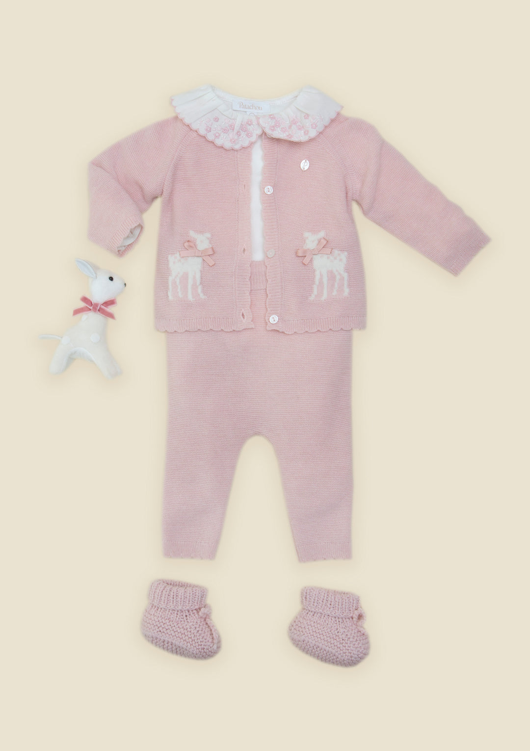 A baby knit outfit set in dusty pink, featuring a long-sleeve cardigan with white deer embroidery, a white ruffled collar, matching pants, booties, and a small white plush toy, all laid out on a light beige background.