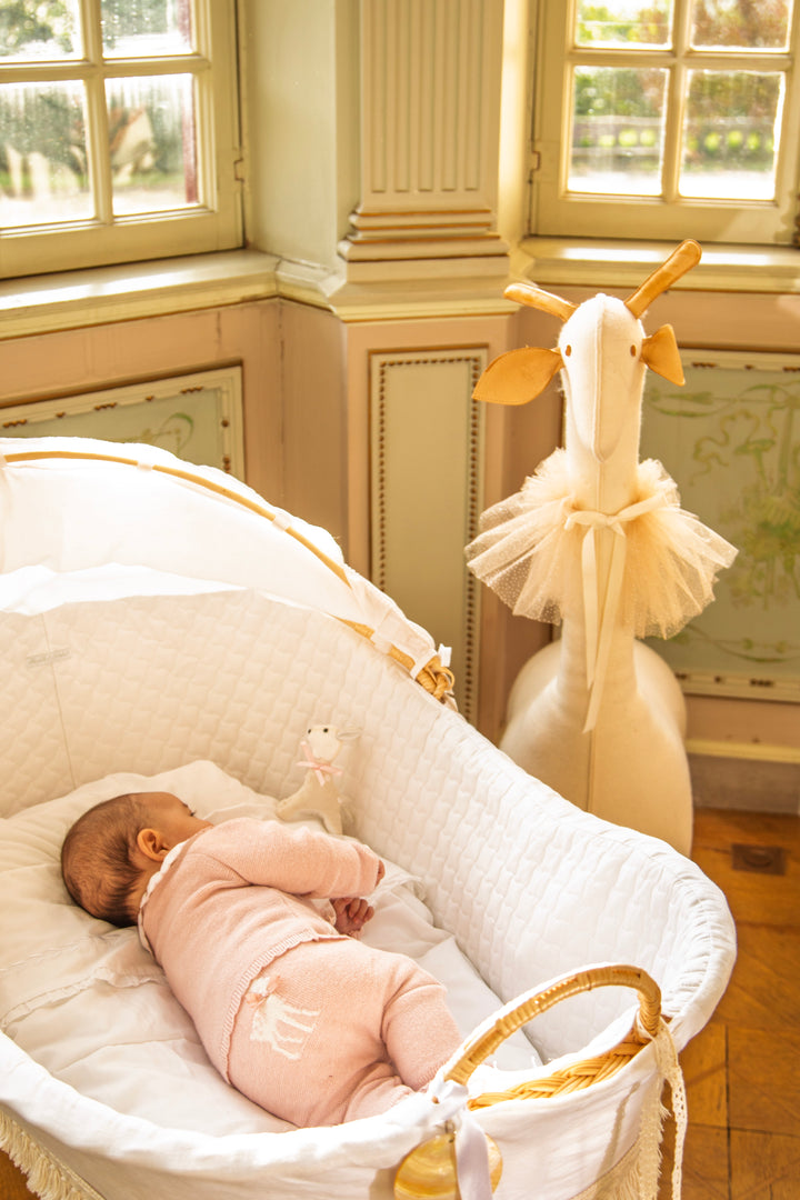 A baby in a light pink onesie sleeping in a white and gold woven bassinet, set in a beautifully decorated nursery with warm sunlight and a decorative giraffe toy.