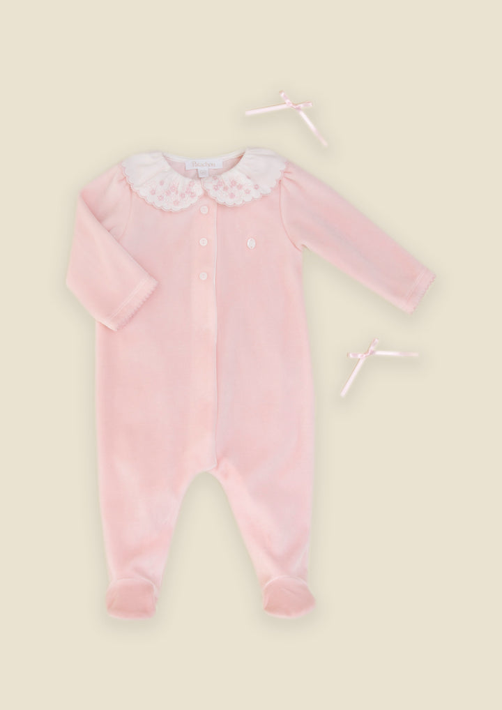 A baby long-sleeve footed sleeper in light pink velvet with a white lace Peter Pan collar and two matching white ribbon bows, displayed on a solid pale beige background.
