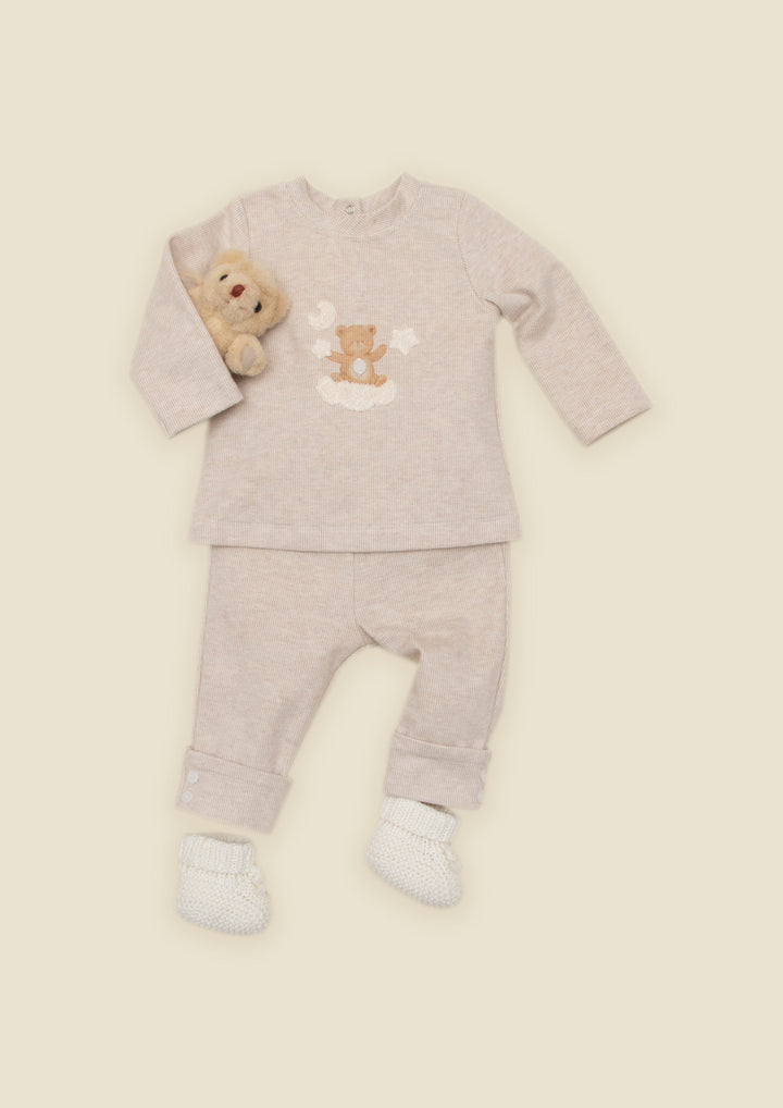 A baby two-piece pajama set in a light heathered beige color, featuring a long-sleeve top with a teddy bear graphic and matching pants, styled with a small plush teddy bear and white booties on a solid cream background.