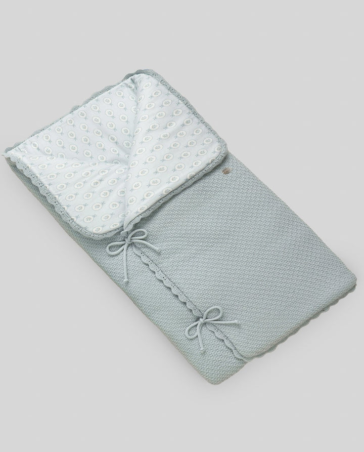 A folded baby blanket or swaddle in a pale sage green color, featuring a textured pattern, a white dotted inner lining, and decorative tie bows, shown on a solid gray background.