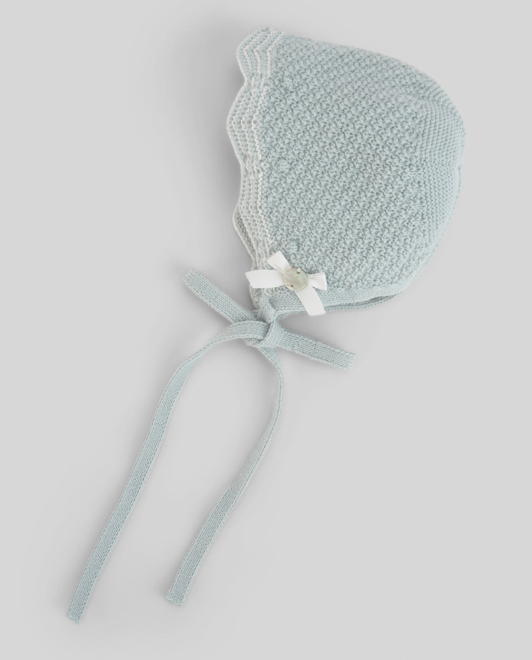 A pale green knit bonnet with a white lace trim, a small white bow, and long ties, shown on a neutral light gray background.