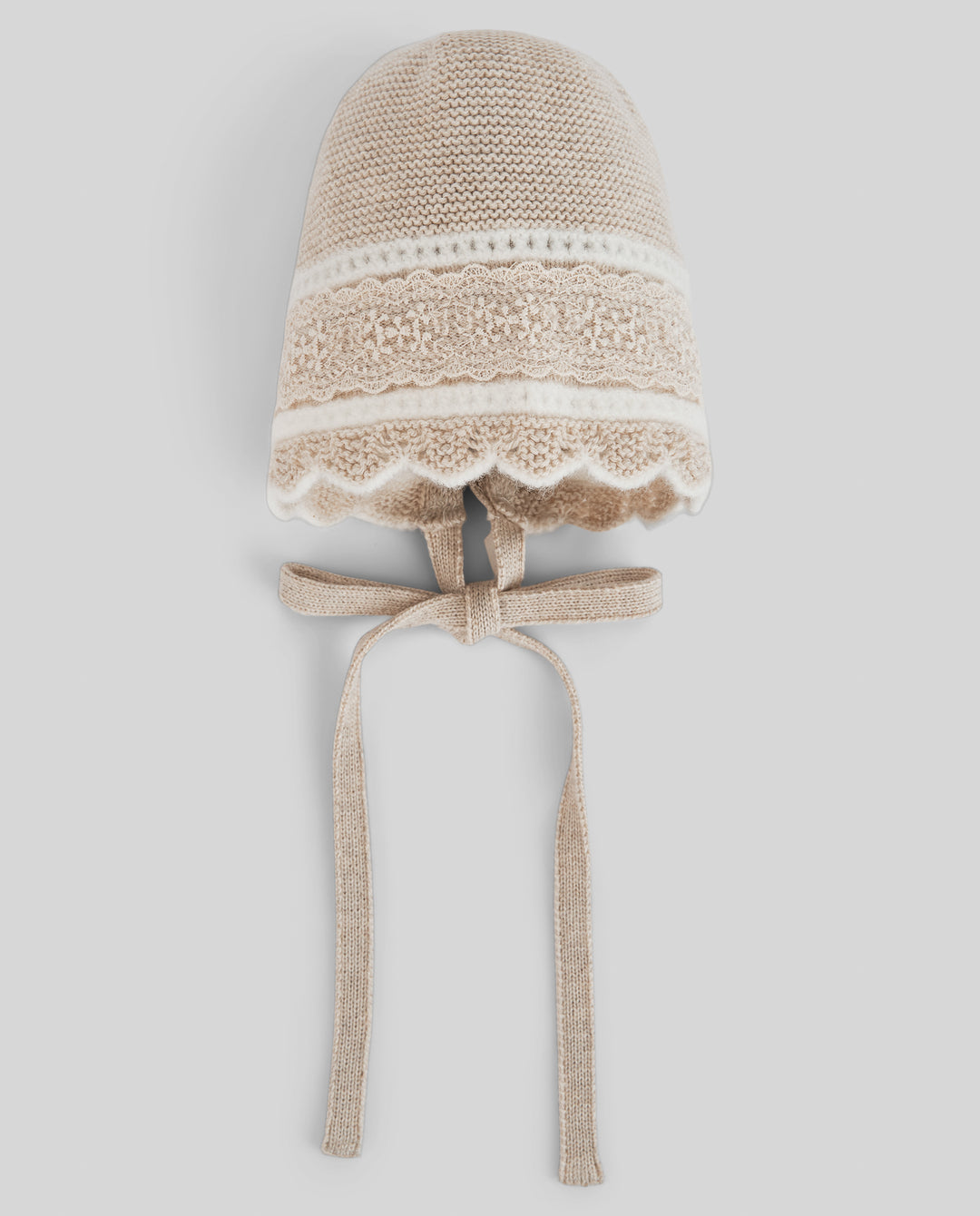 A beige knit bonnet hat with a white lace band, scalloped trim, and matching ribbon ties, shown against a light gray background.