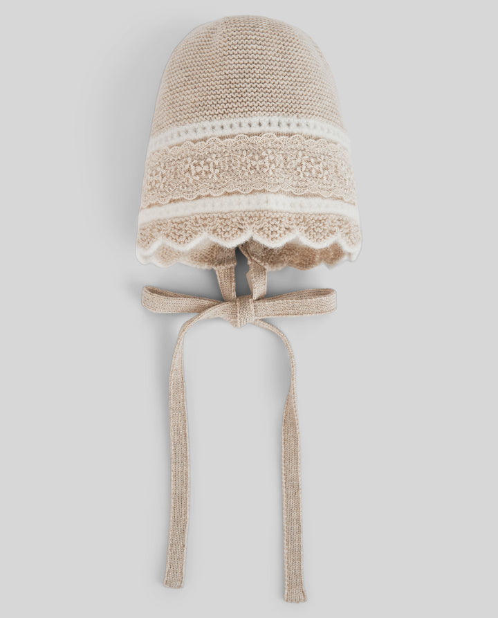A beige knit bonnet hat with a white lace band, scalloped trim, and matching ribbon ties, shown against a light gray background.