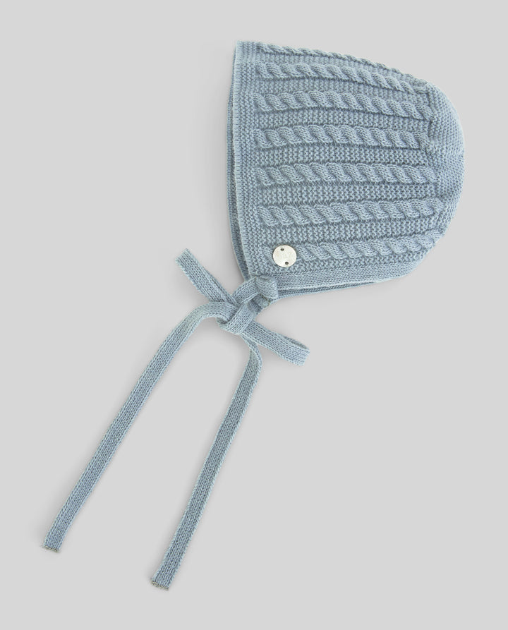 A light blue cable-knit bonnet with tie straps and a small white button, shown on a neutral gray background.
