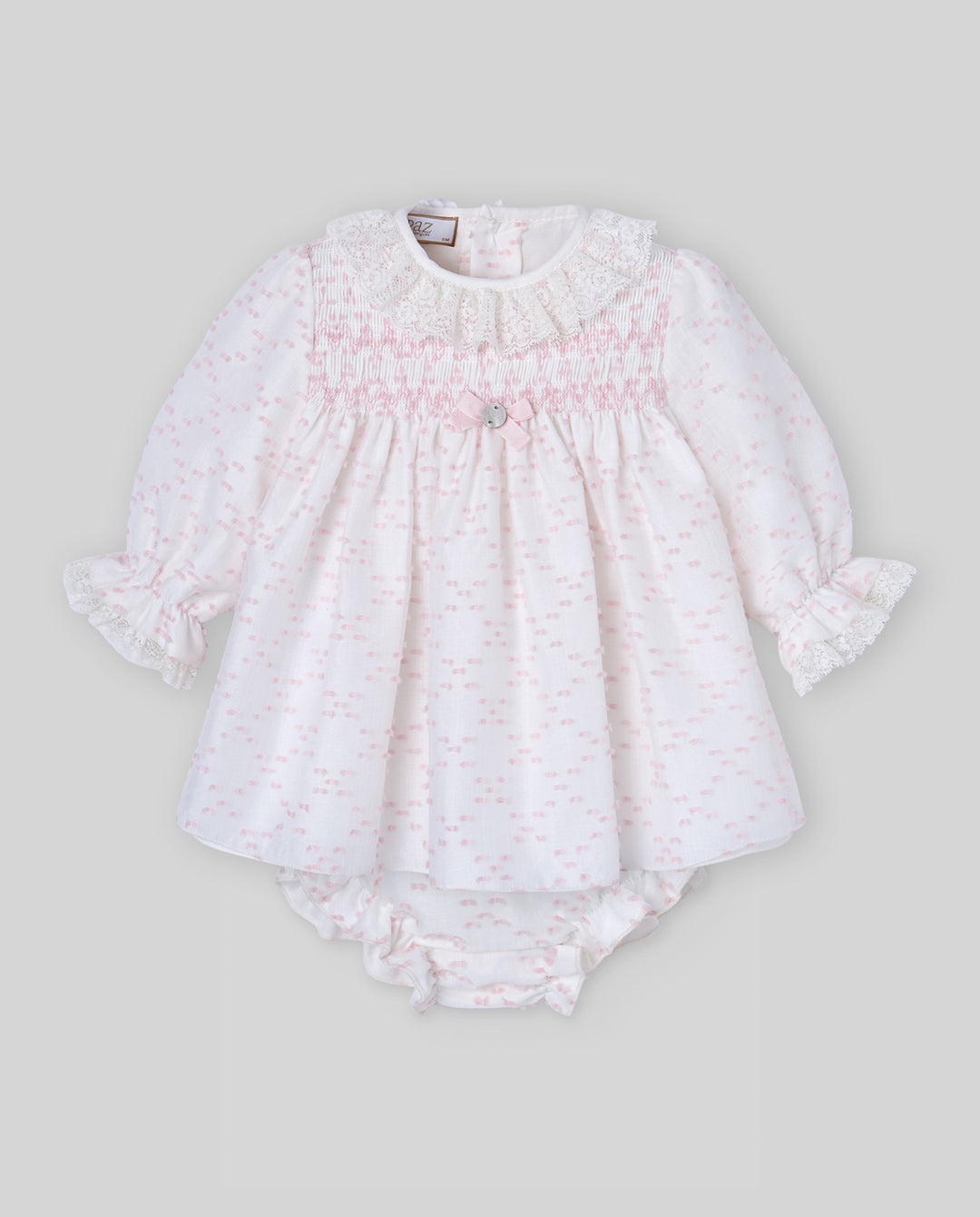 A baby girl's long-sleeve smocked romper in white fabric with a delicate light pink pattern, featuring ruffled cuffs and a matching ruffled diaper cover, shown against a solid gray background.