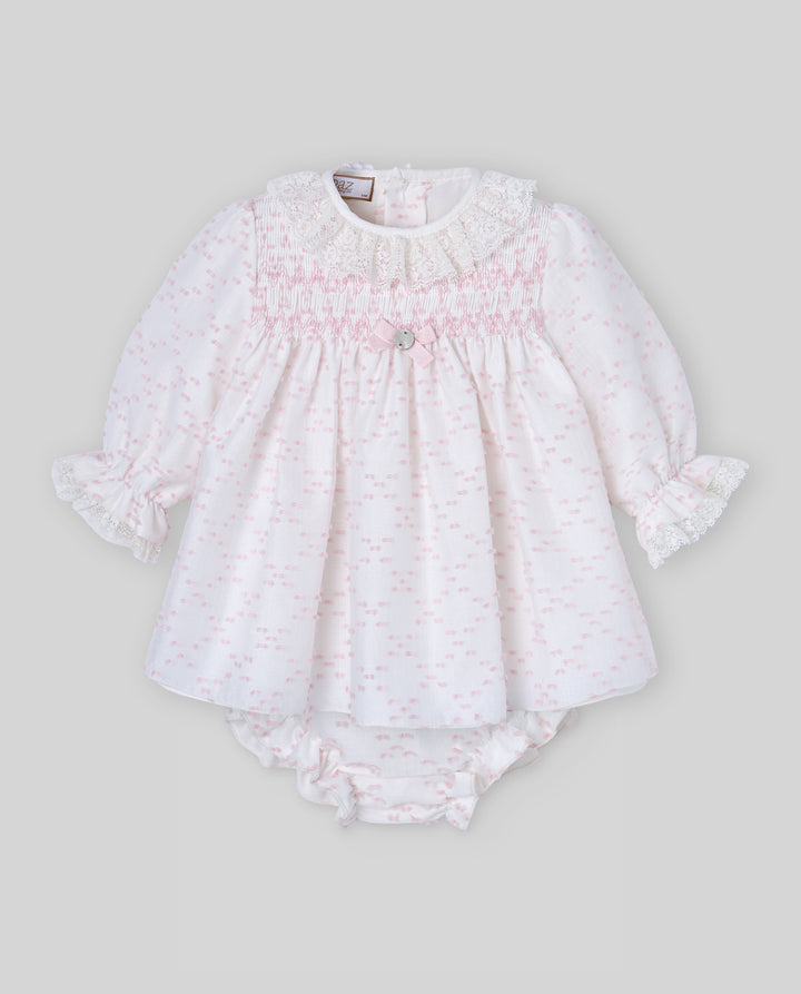A baby girl's long-sleeve smocked romper in white fabric with a delicate light pink pattern, featuring ruffled cuffs and a matching ruffled diaper cover, shown against a solid gray background.