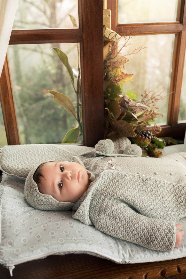 A baby wearing a light gray knit bonnet and matching textured knit outfit lies on a patterned blanket in front of a wooden window with soft natural light.