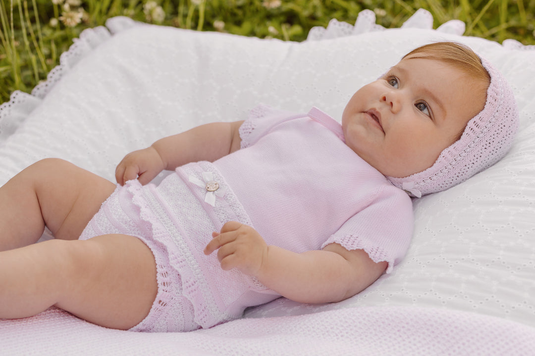 Baby in pink outfit and hat lying on a white blanket with grass in the background