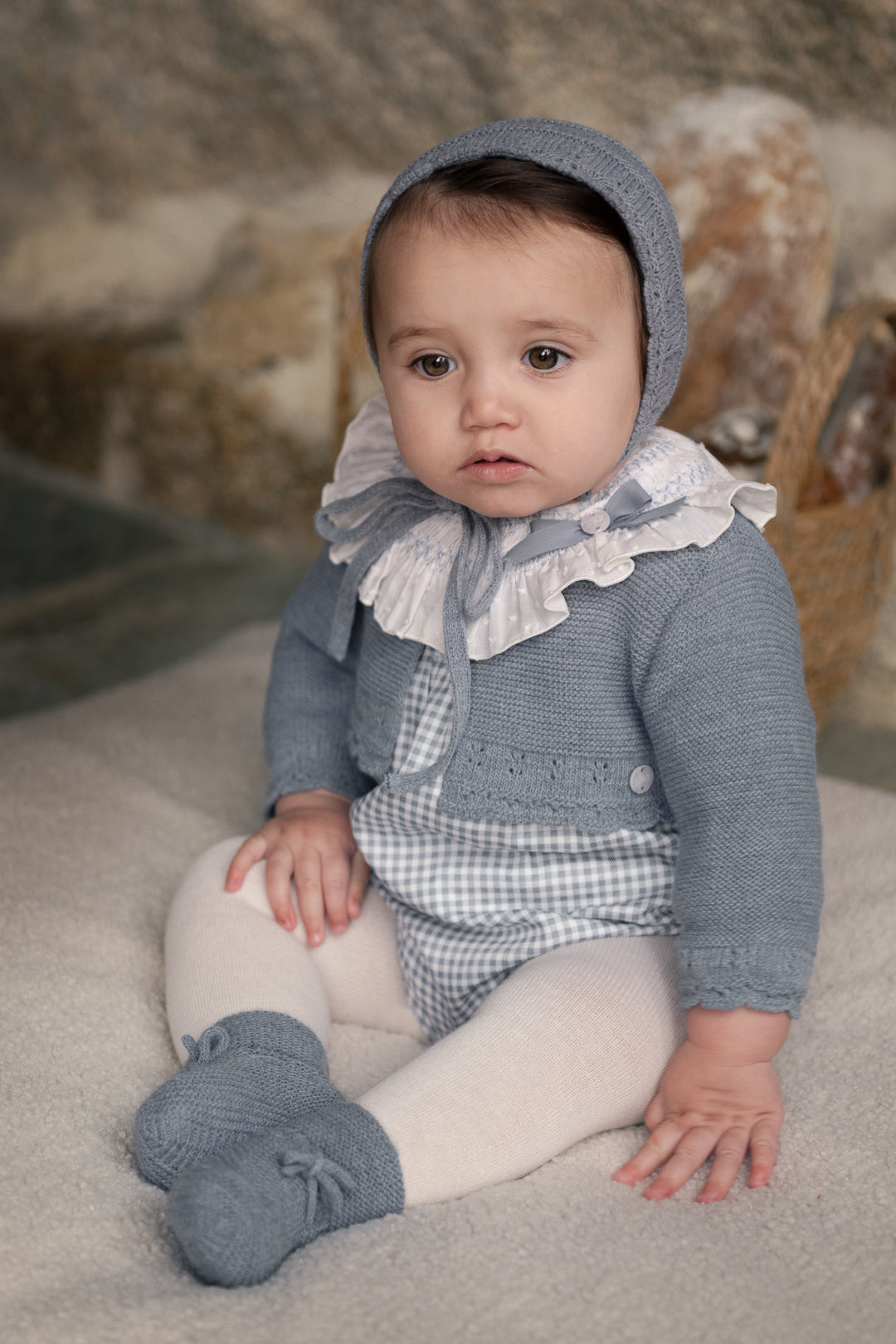 A baby girl wearing a blue-gray knit bonnet, matching booties, a blue-gray cardigan, and a blue-and-white gingham romper with a white ruffled collar, sitting on a soft beige rug.