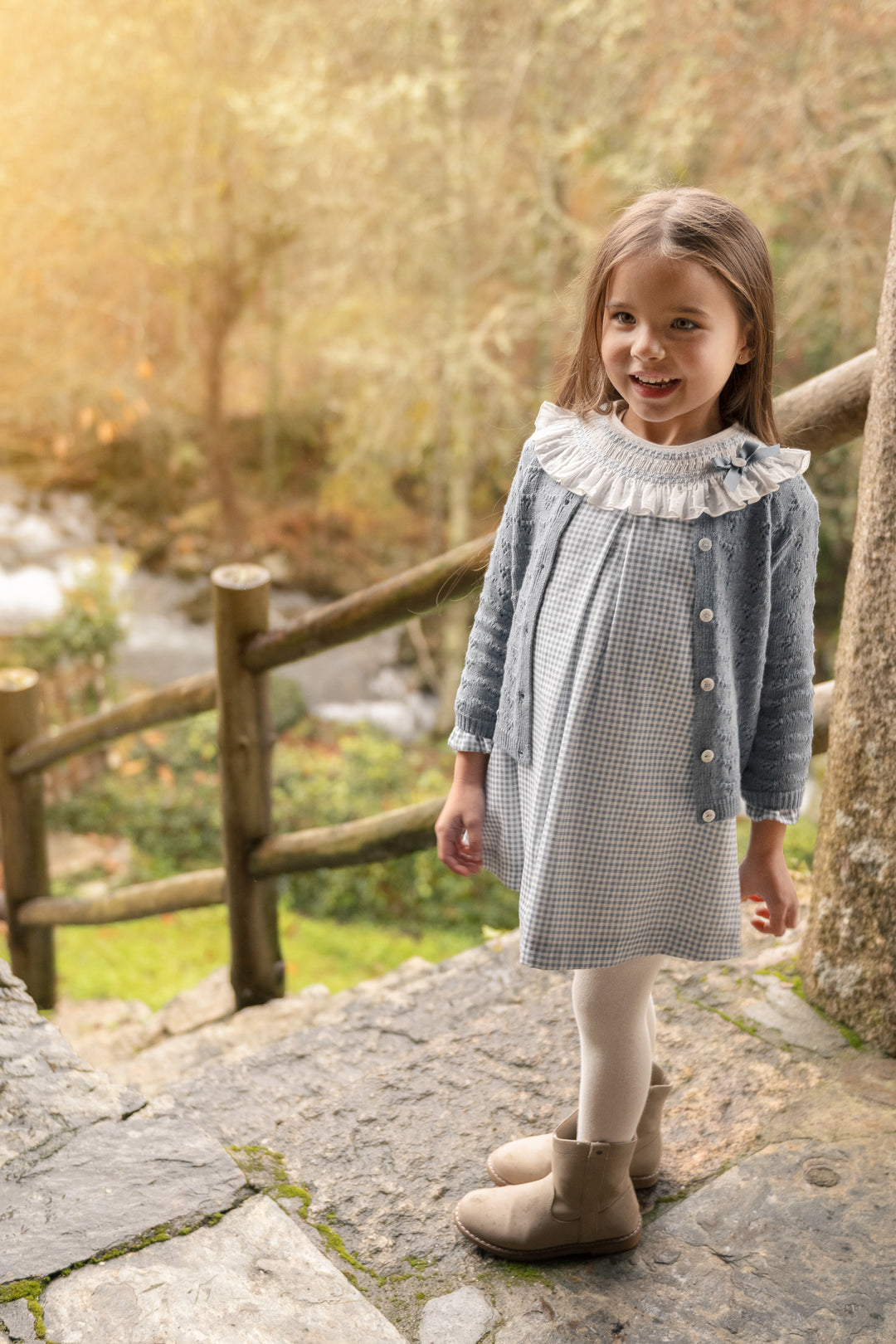 A young girl stands on stone steps outdoors wearing a light gray and white gingham dress with a ruffled white collar, a blue-gray button-front cardigan, white tights, and beige ankle boots. A wooden fence and blurred trees are in the background.